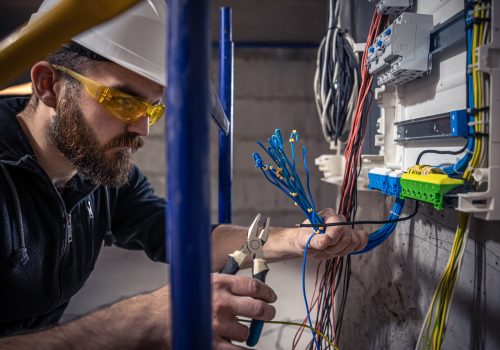 A male electrician works in a switchboard with an electrical connecting cable, connects the equipment with tools.